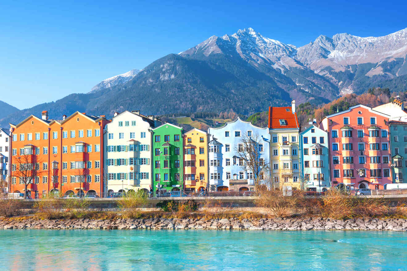 Colorful facades of historical buildings along the Inn River in Innsbruck, with snow-capped mountains in the backdrop under a clear blue sky